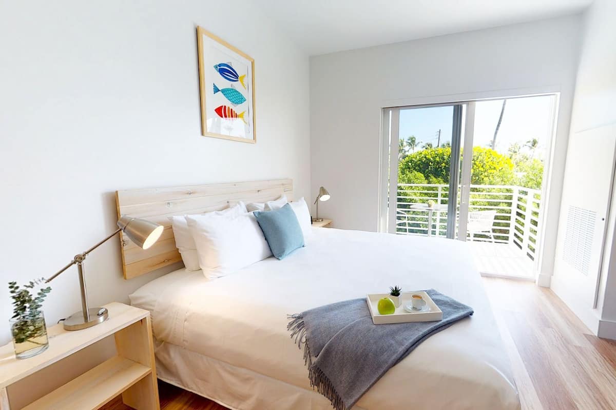 A bedroom with a large bed, a nightstand, a bedside table, a framed picture, and a balcony with a view of greenery.