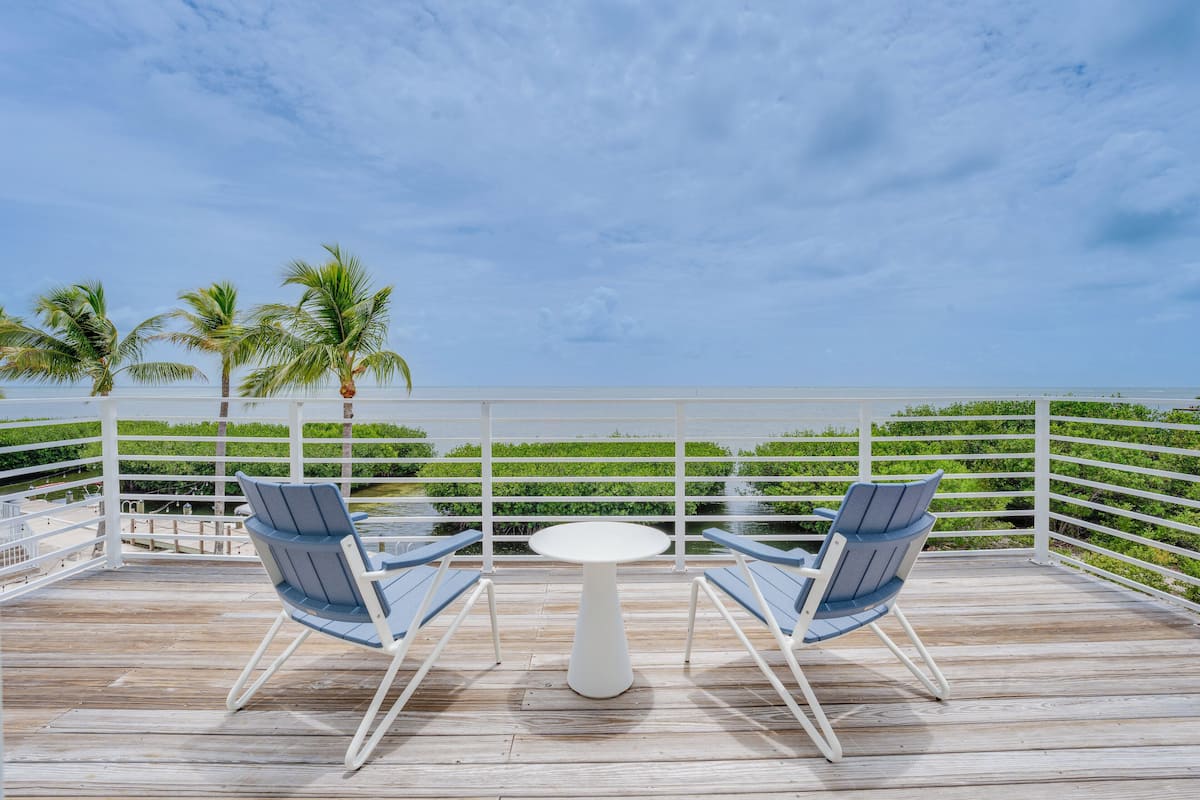 A deck with two blue chairs and a white table, overlooking a body of water and palm trees.