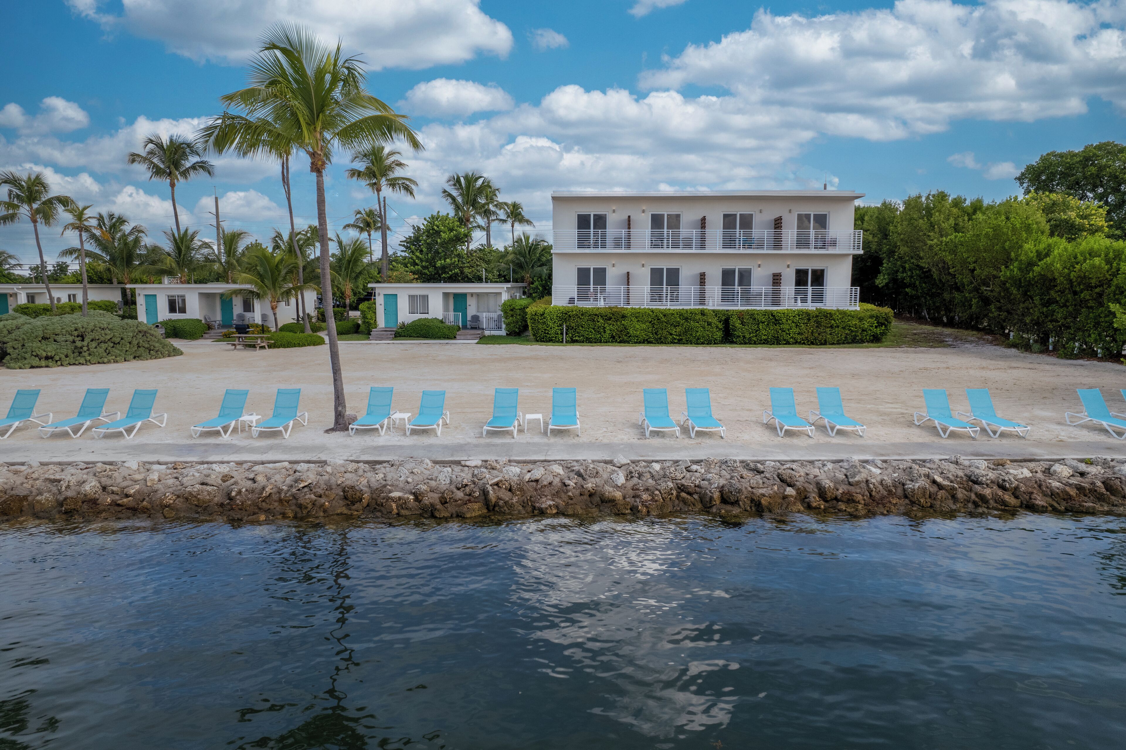 Hotel frente a la playa con zona de piscina, palmeras y un edificio de dos pisos.