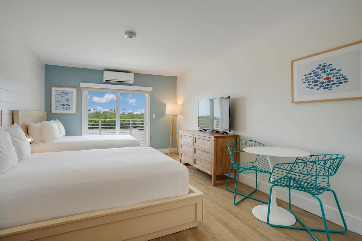 A hotel room with two beds, a desk with a TV, teal chairs, and a balcony with a view.
