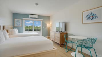 A hotel room with two beds, a desk with a TV, teal chairs, and a balcony with a view.