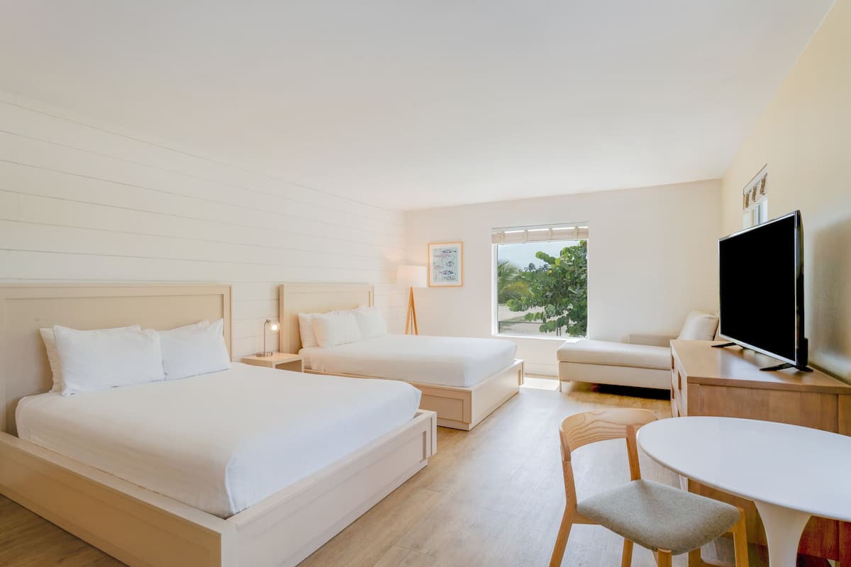 A hotel room with two beds, a TV, a desk, and a window with a view of greenery.