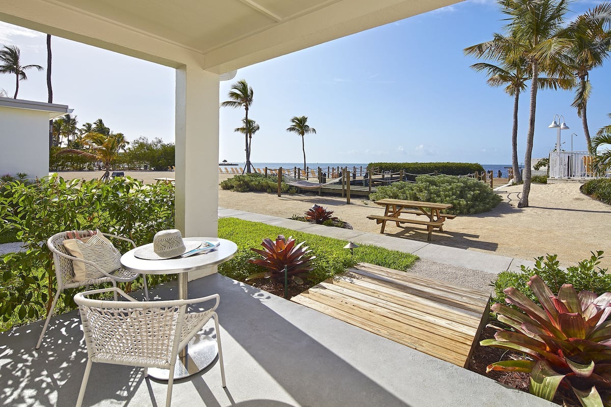 A covered outdoor seating area with wicker chairs and a table, overlooking a beach with palm trees and a wooden deck.