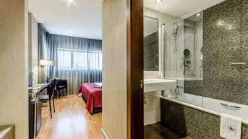 Habitación de hotel con suelo de madera, un escritorio con silla, cama con colcha roja, baño con espejo grande, bañera y ducha.