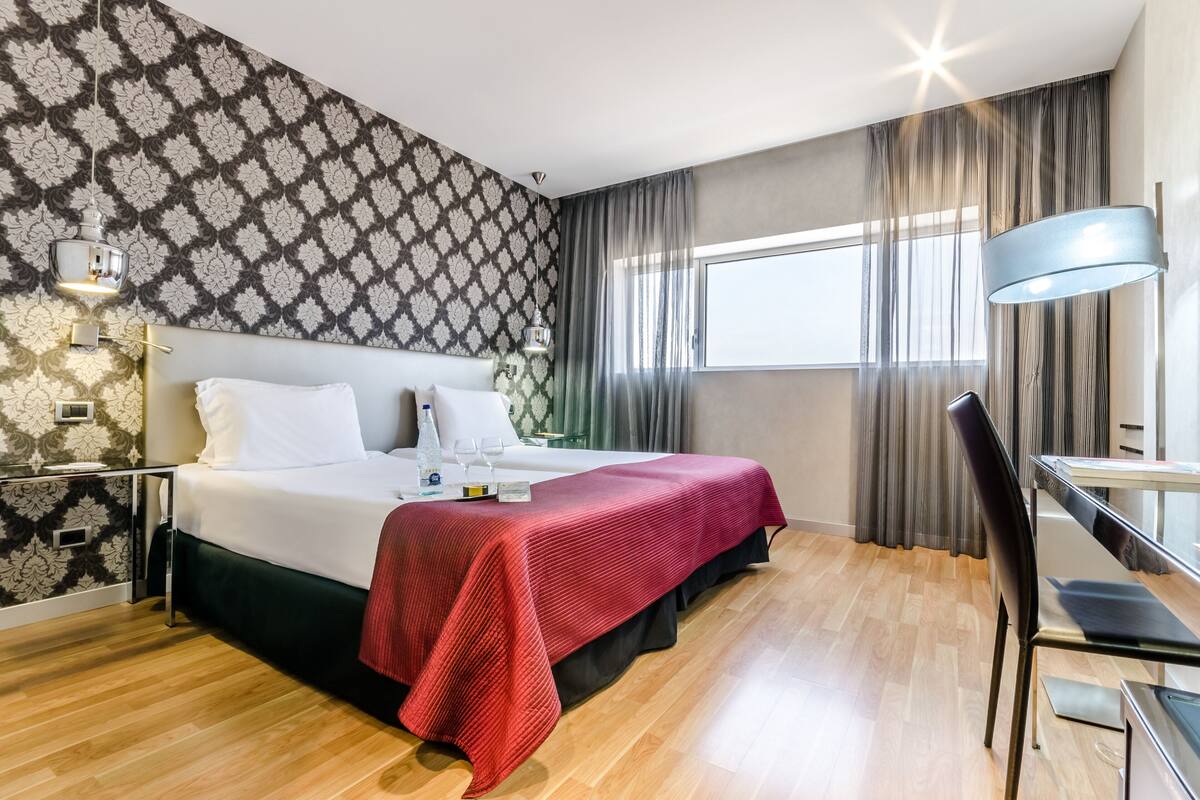 Una habitación de hotel con cama, escritorio, silla, ventana con cortinas y un papel tapiz estampado.
