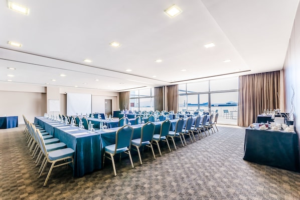 Un salón de conferencias con filas de mesas y sillas, ventanas amplias y suelo alfombrado.