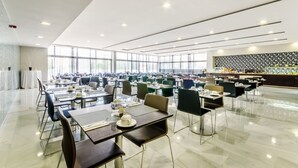 Un vaste espace de salle à manger avec plusieurs tables et chaises, de grandes fenêtres et une cuisine moderne en arrière-plan.