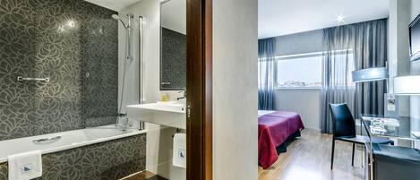 Un baño moderno con ducha, lavamanos y espejo. Se ve una cama con una colcha roja y un escritorio con una silla.