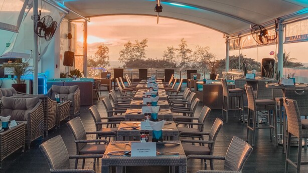 Ein überdachter Außenbereich zum Essen mit Tischen und Stühlen, ein Baldachin darüber und Blick auf den Sonnenuntergang im Hintergrund.