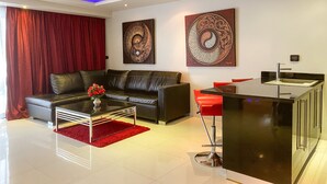 Ein modernes Wohnzimmer mit einer schwarzen Ledersofa, einem Glastisch und roten Akzenten. Zwei Kunstwerke sind an der Wand angebracht, und es gibt eine Küchenzeile mit Spüle und roten Barhocker.