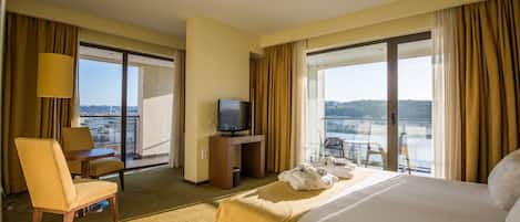 Habitación de hotel con cama, escritorio con televisor, una silla y vistas a la ciudad a través de amplias ventanas.