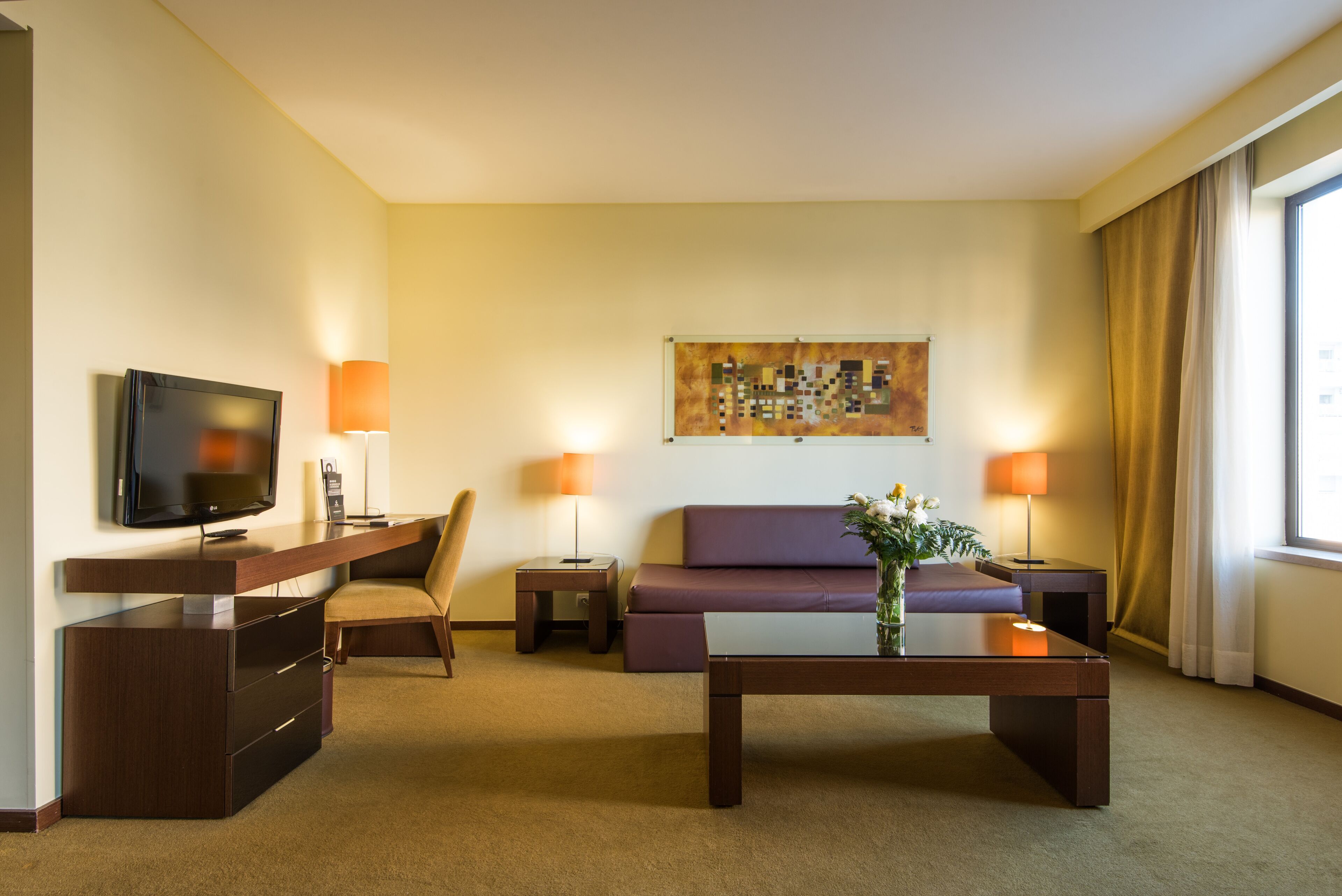 Hotelkamer met een flatscreen televisie, een bank, een bijzettafeltje, twee lampen, een schilderij aan de muur en een raam met gordijnen.