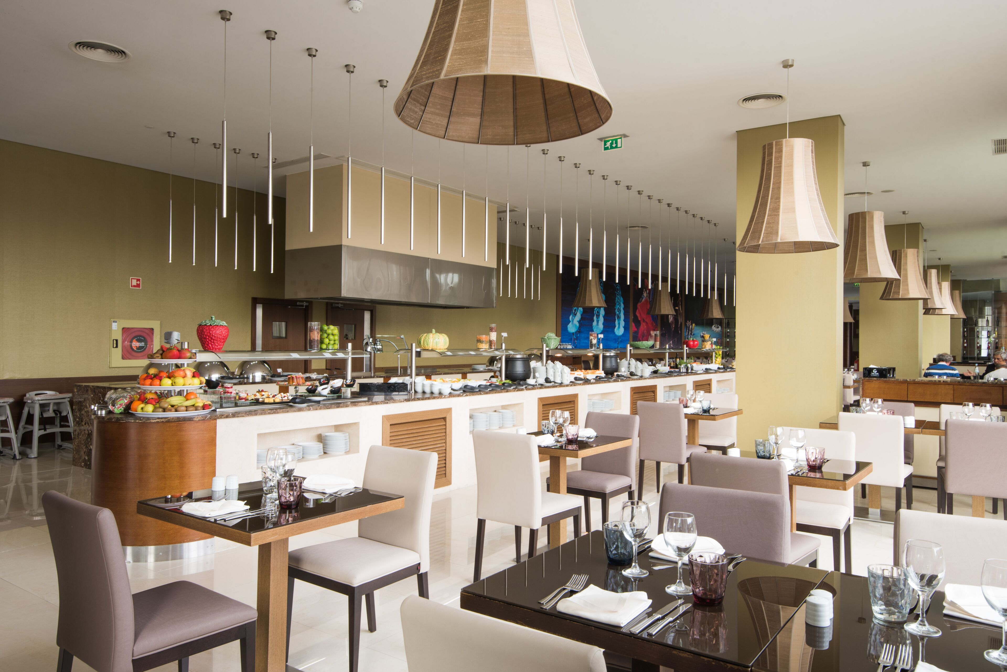 A modern restaurant with multiple dining tables and chairs, a central bar area with a fruit display, and pendant lighting.