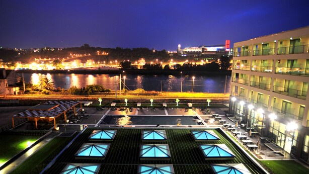 A hotel with a rooftop terrace featuring a pool and outdoor seating, overlooking a body of water and a cityscape at night.