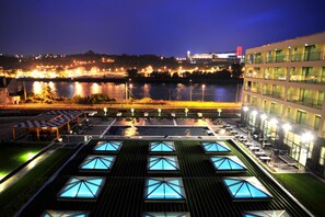 Un hotel con terraza en la azotea que cuenta con piscina y mobiliario exterior, con vistas a un cuerpo de agua y un paisaje urbano nocturno.