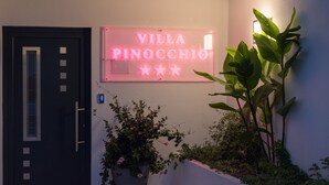 Entrada de un edificio con un letrero de neón que dice "Villa Pinocchio" y una planta en maceta.