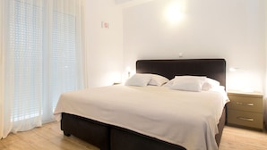 Una cama bien hecha con un cabecero oscuro, una mesita de noche con lámpara y una ventana con cortinas de gasa.