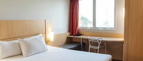Una habitación de hotel con cama, escritorio, silla y ventana con cortinas.
