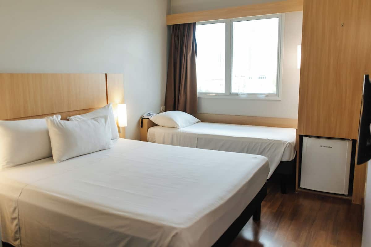 A hotel room with two beds, a small refrigerator, a desk with a lamp, and a window with curtains.