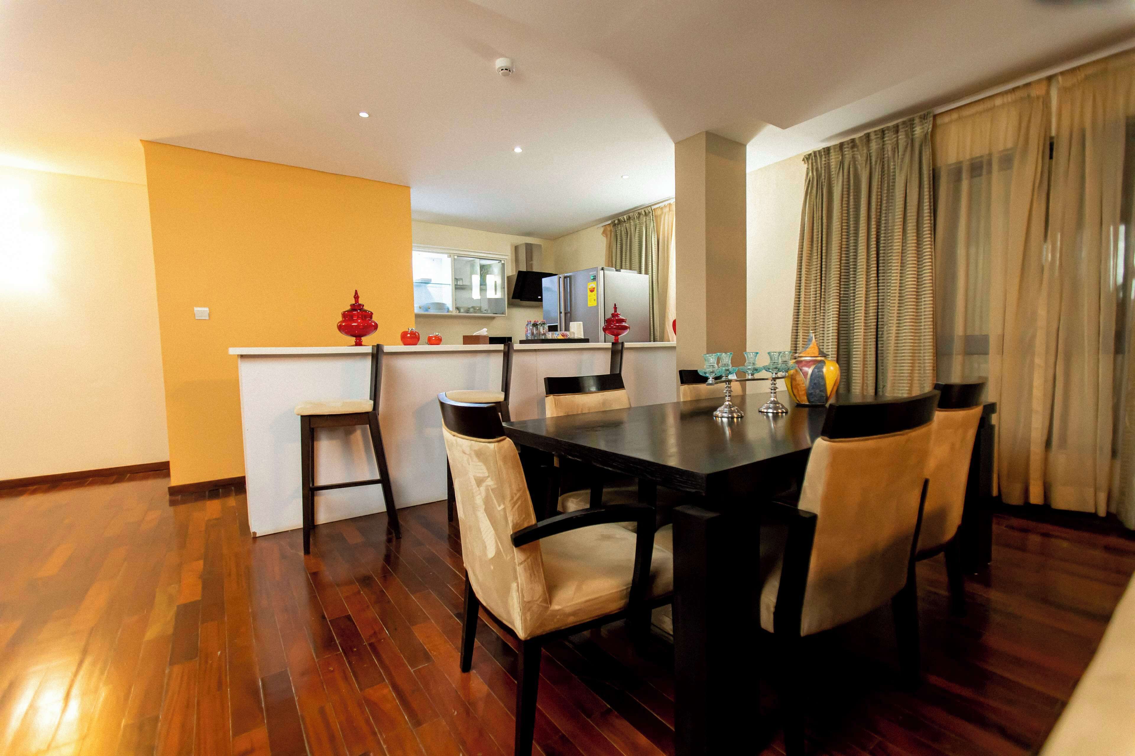 A dining area with a wooden table and chairs, a kitchen in the background, and a yellow wall.