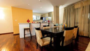 A dining area with a wooden table and chairs, a kitchen in the background, and a yellow wall.