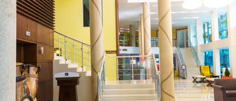 A spacious lobby with a central staircase, a reception desk, and a seating area with a yellow chair.
