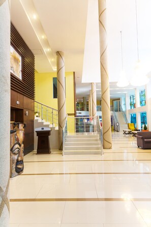 Un hall d’entrée spacieux avec un escalier central, un comptoir d’accueil et un coin salon agrémenté d’un fauteuil jaune.