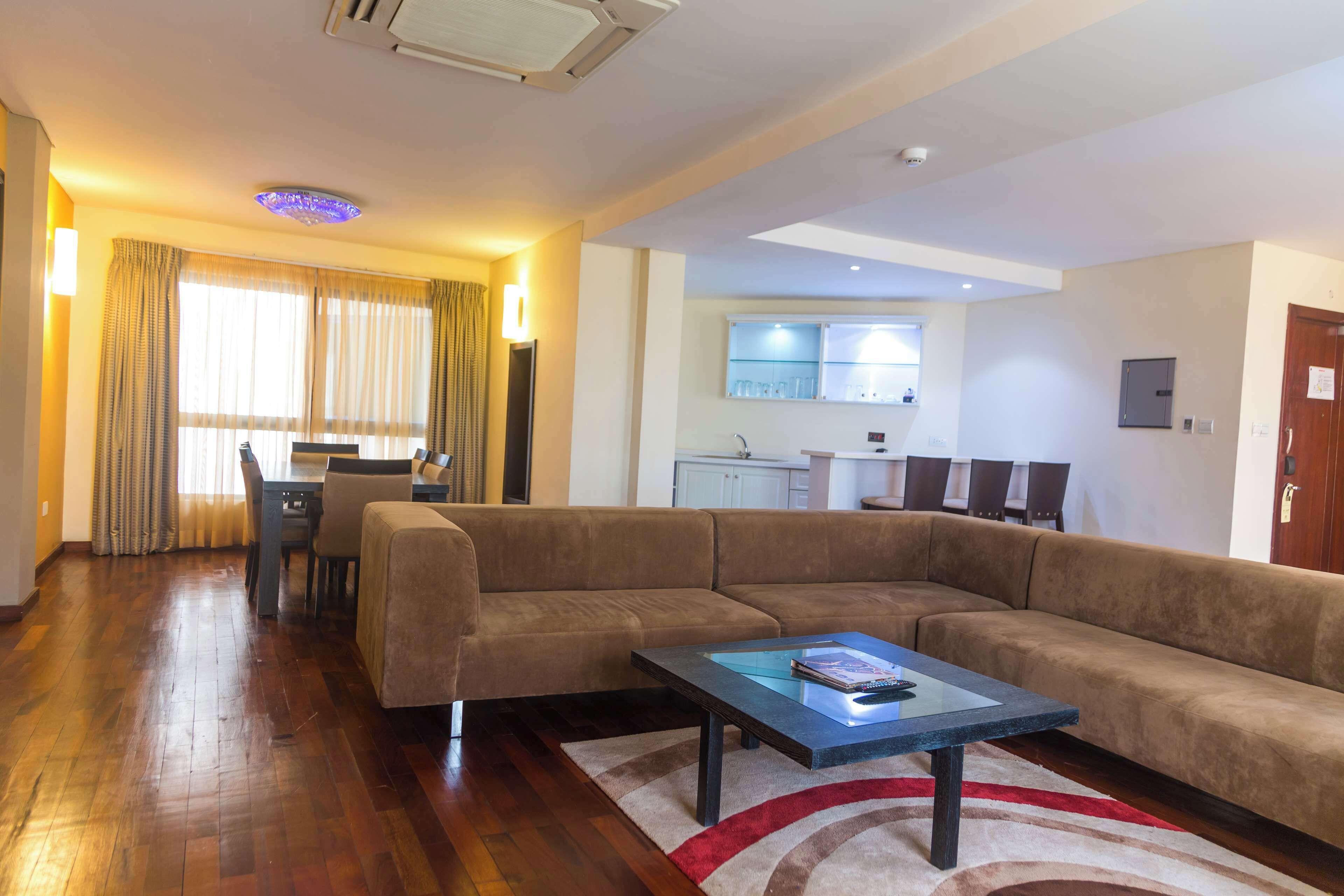 A spacious living area with a large brown sofa, a dining table, and a kitchen in the background.