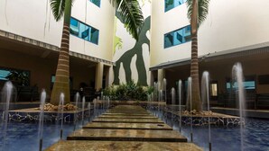 A pool area with a central water feature and palm trees, surrounded by a building with large windows.