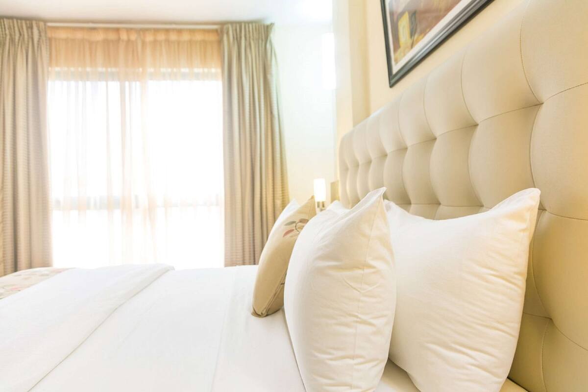 Habitación de hotel con una cama grande, cortinas color beige y un cabecero capitonado.