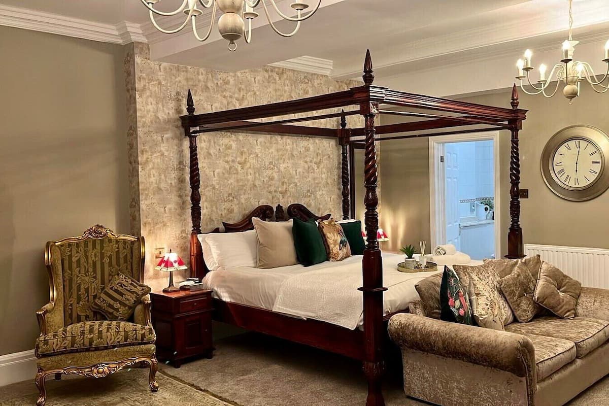 A four-poster bed with a wooden frame, a plush armchair, a small table with a lamp, and a wall clock.