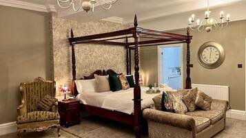 A four-poster bed with a wooden frame, a plush armchair, a small table with a lamp, and a wall clock.