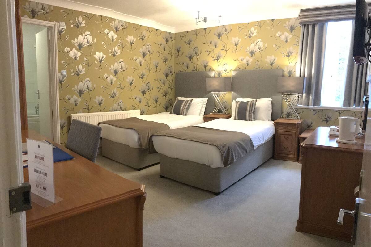 A hotel room with two beds, a desk, a chair, a radiator, a window with curtains, and a floral wallpaper.