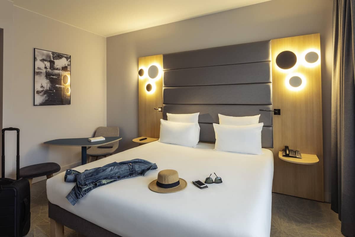 Una habitación de hotel con cama, mesitas de noche, una silla, una maleta y un sombrero sobre la cama.