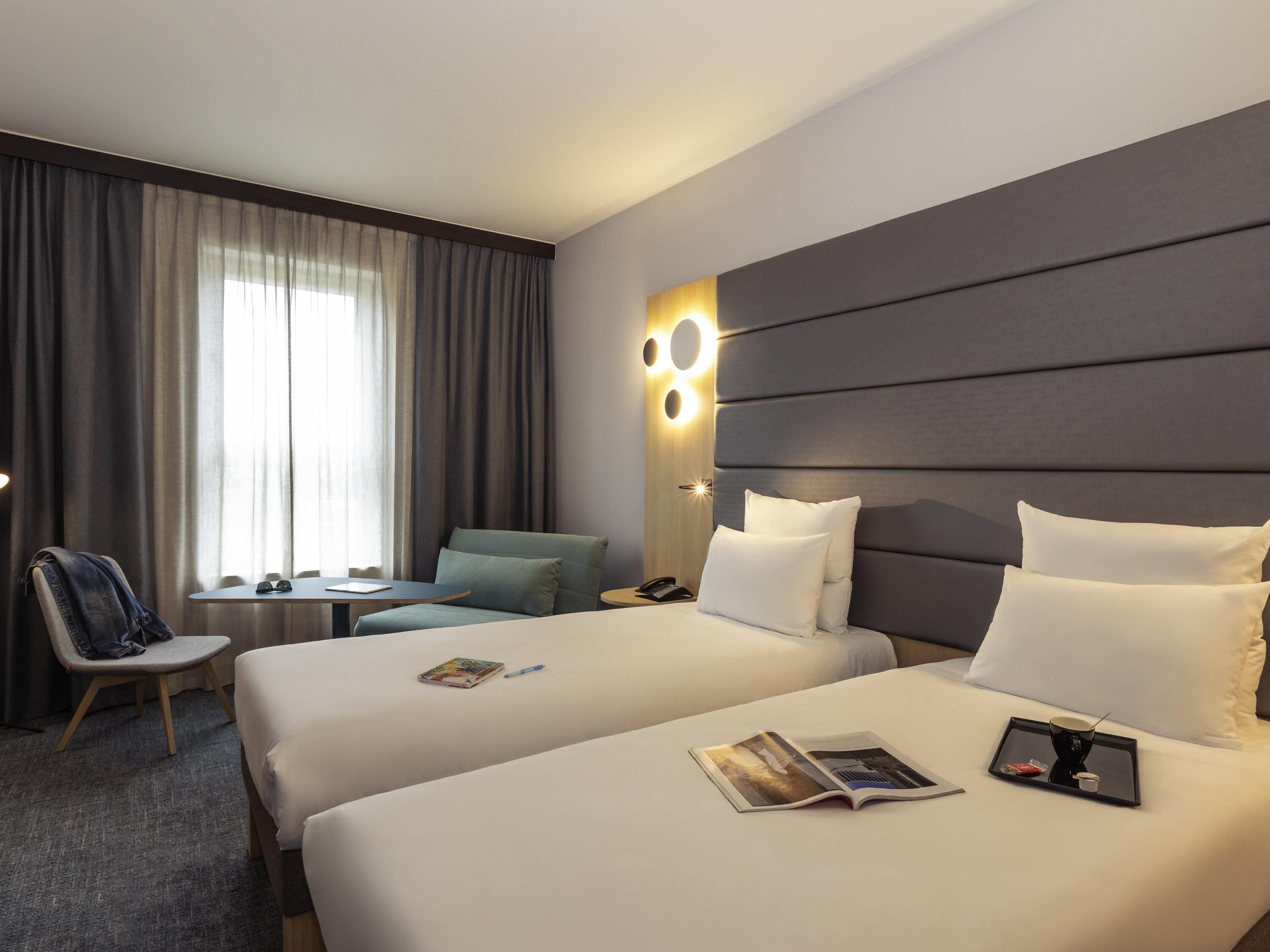 A hotel room with two beds, a desk, a chair, and a large window with curtains.
