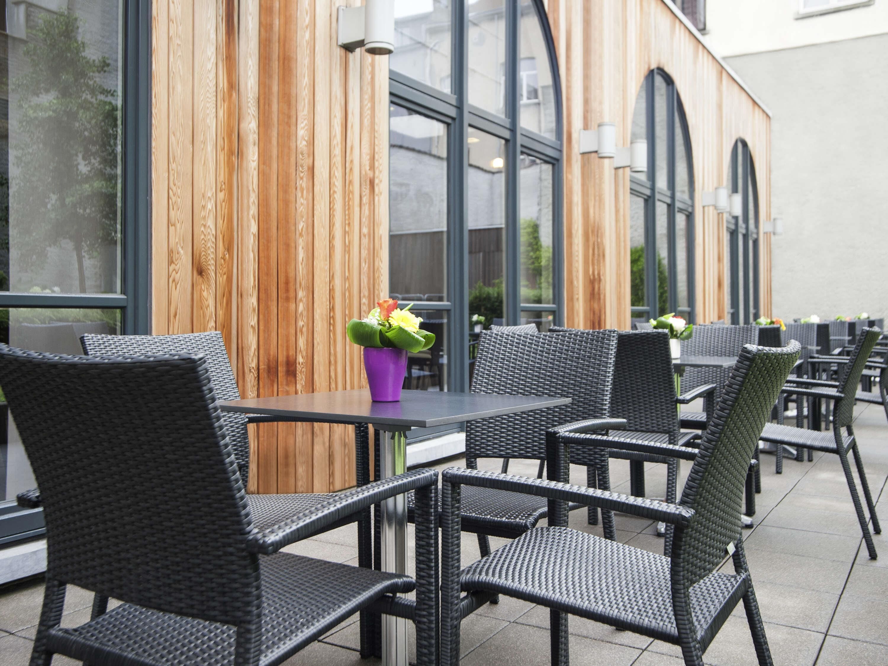 Un espace extérieur aménagé avec des chaises et des tables en osier, un mur en bois et de grandes fenêtres.