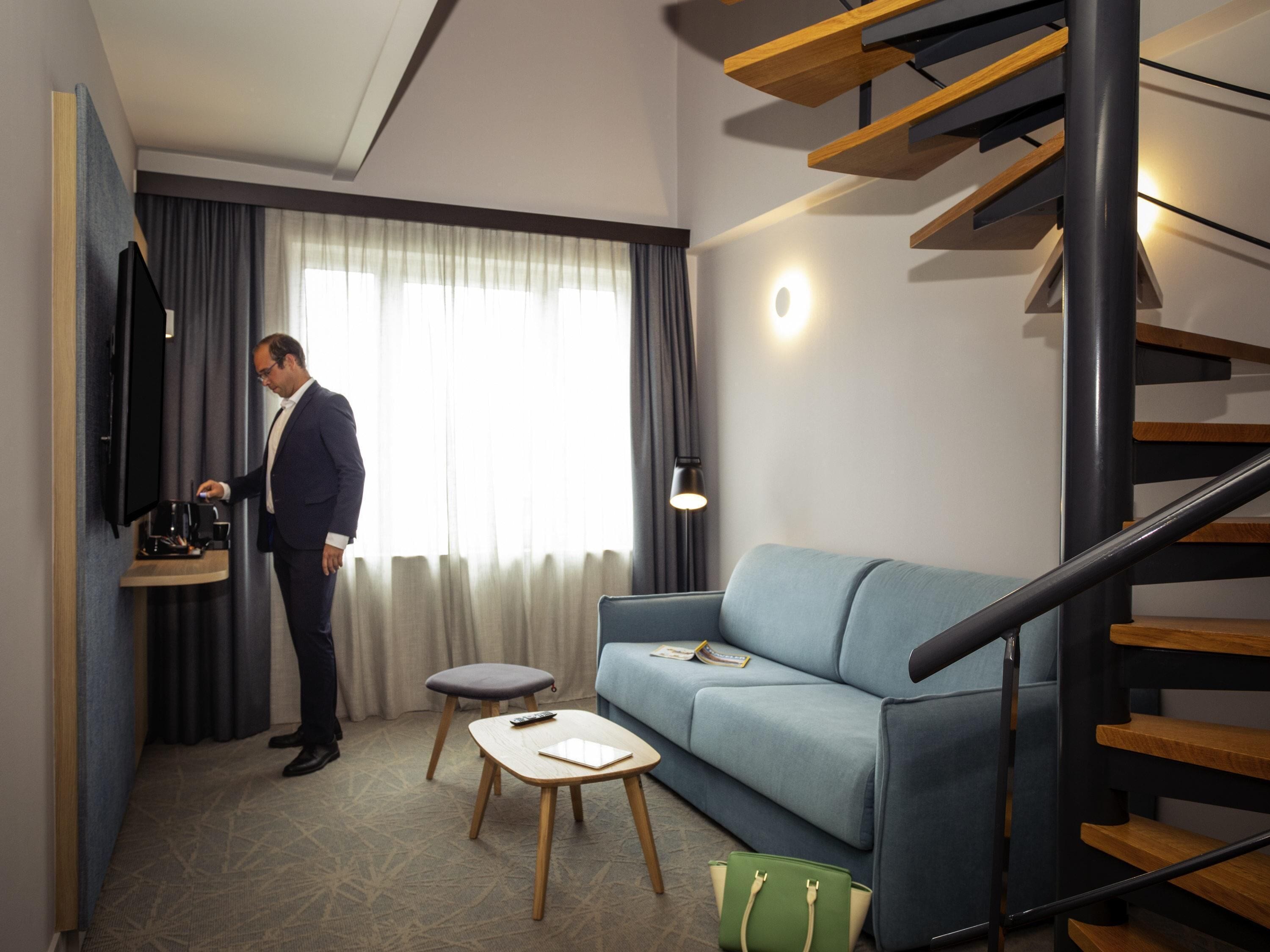 A modern hotel room with a sofa, a small table, a coffee machine, a staircase, and a window with curtains.