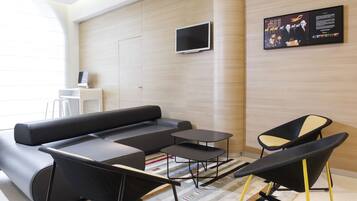 A modern hotel room with a sectional sofa, two black and yellow chairs, a flat-screen TV, and a wall-mounted information board.
