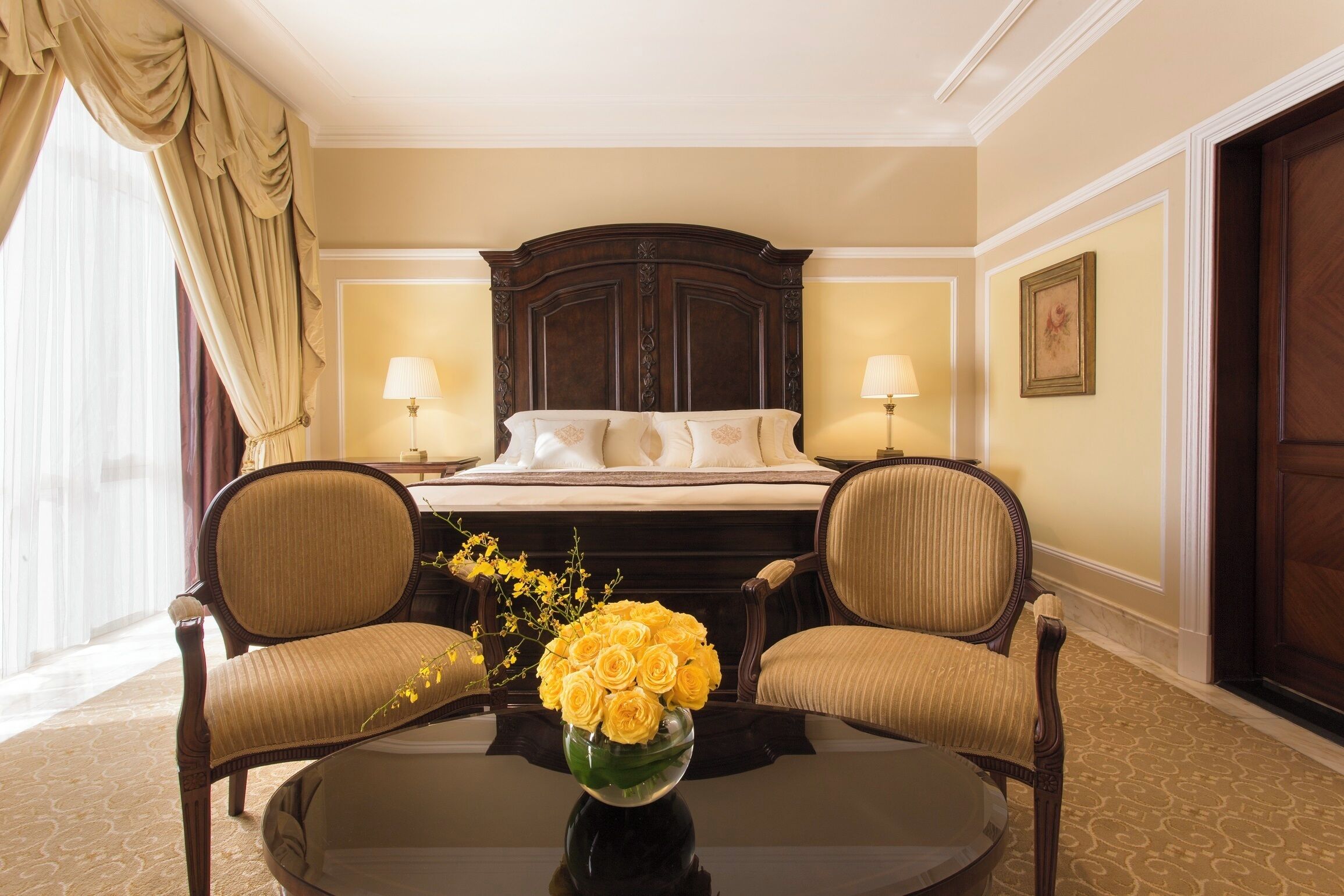 Habitación de hotel con una cama grande, dos sillones, una mesa de centro redonda con un jarrón de flores amarillas y un ventanal con cortinas.