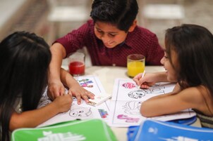 Tres niños participan en una actividad de dibujo en una mesa, con material de colorear y un jugo.