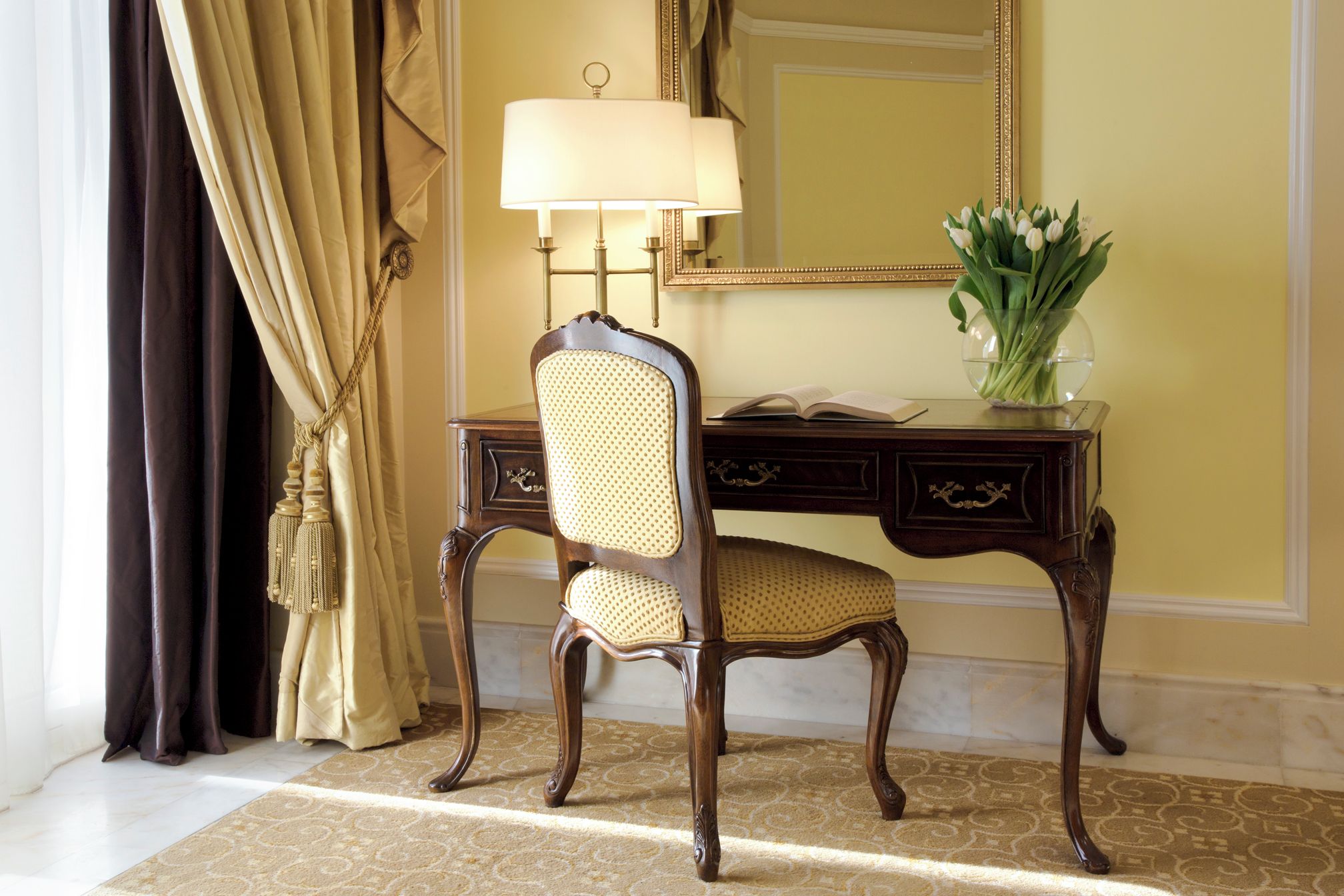 Una habitación con un escritorio de madera, una silla, una lámpara, un espejo y un jarrón con flores.