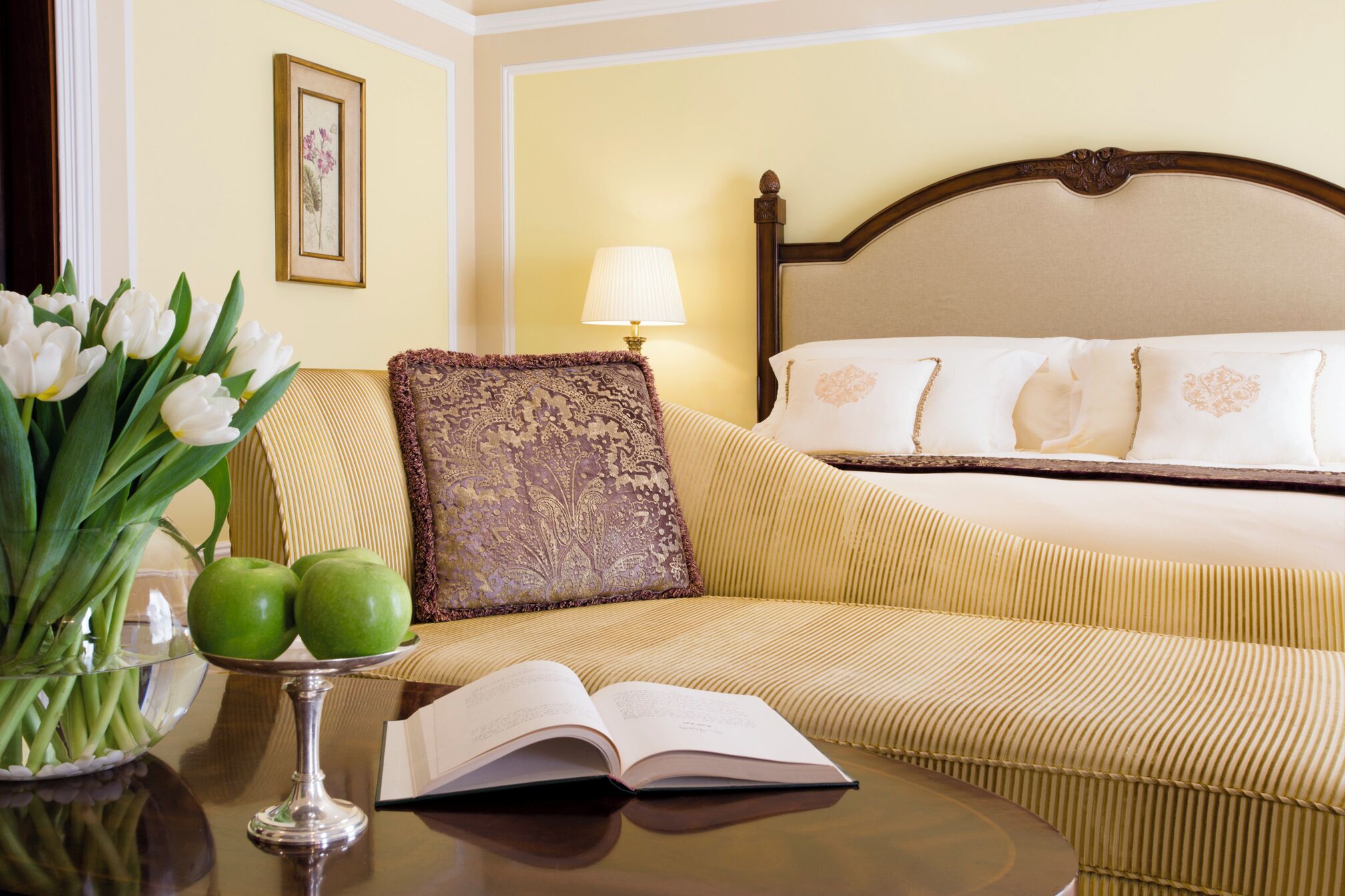 A hotel room with a bed, a bedside table with a book and a glass of water, a vase with flowers, and a lamp.