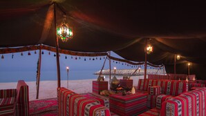 A tented area with patterned sofas and cushions, a fire pit, and hanging lanterns. - The Regency Hotel, Kuwait (Salmiya)