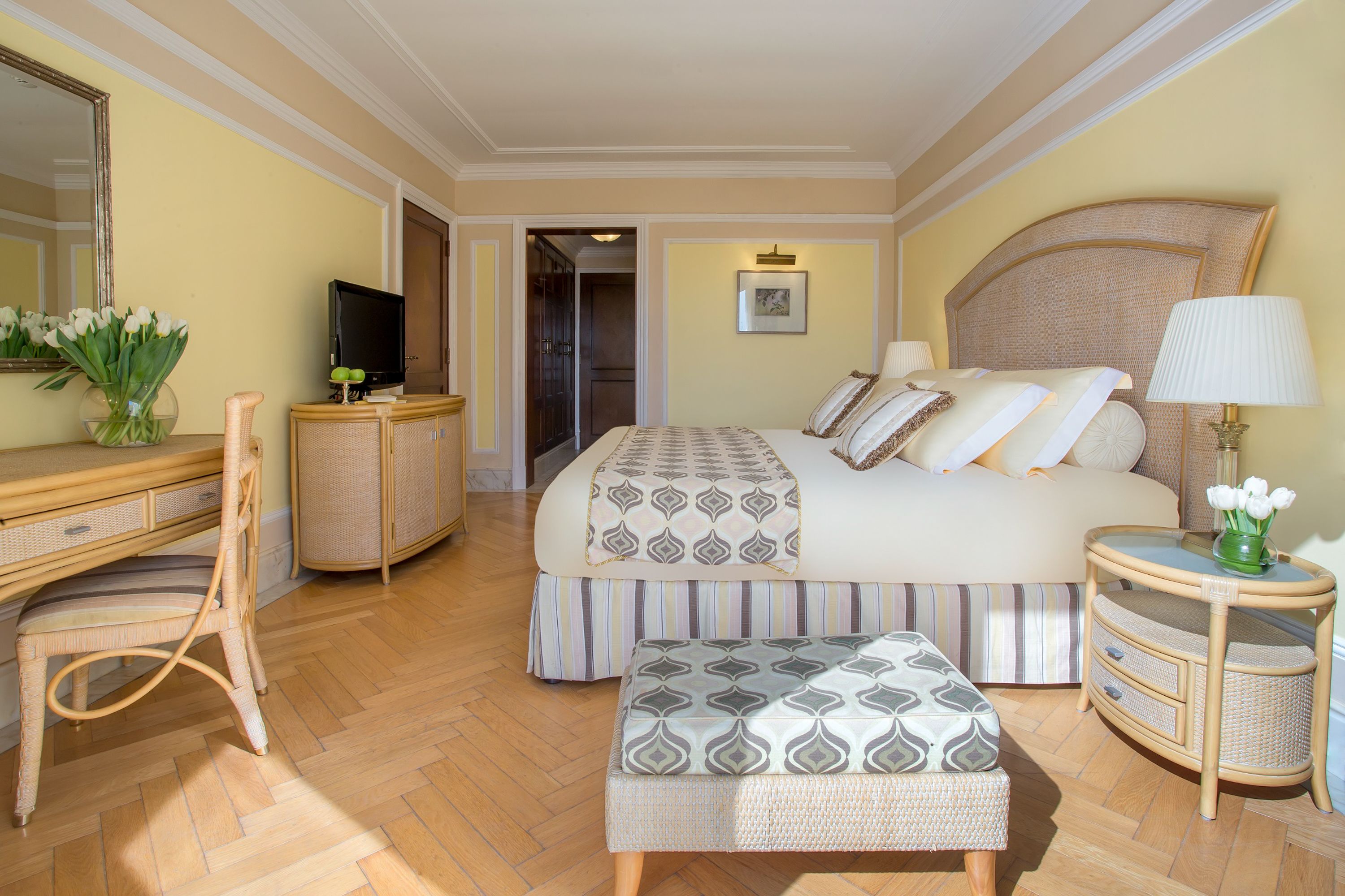 Una habitación de hotel con una cama grande, un escritorio con televisor, una silla, una mesita de noche con lámpara y un jarrón con flores.