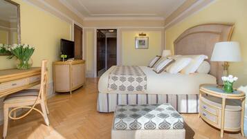 Una habitación de hotel con una cama grande, un escritorio con televisor, una silla, una mesita de noche con lámpara y un jarrón con flores.