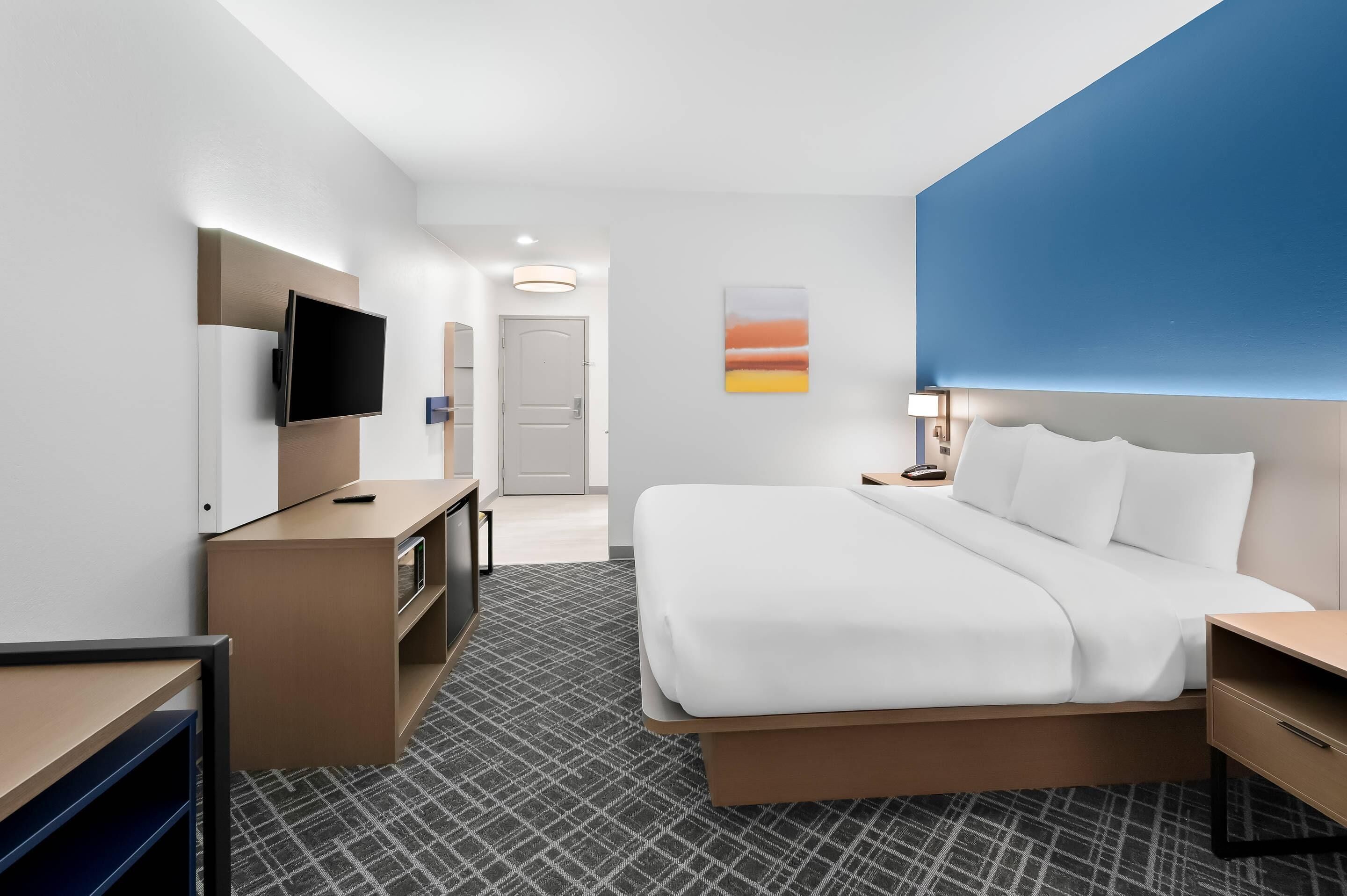A hotel room with a large bed, a desk with a flat-screen TV, a small seating area, and a door leading to a bathroom.
