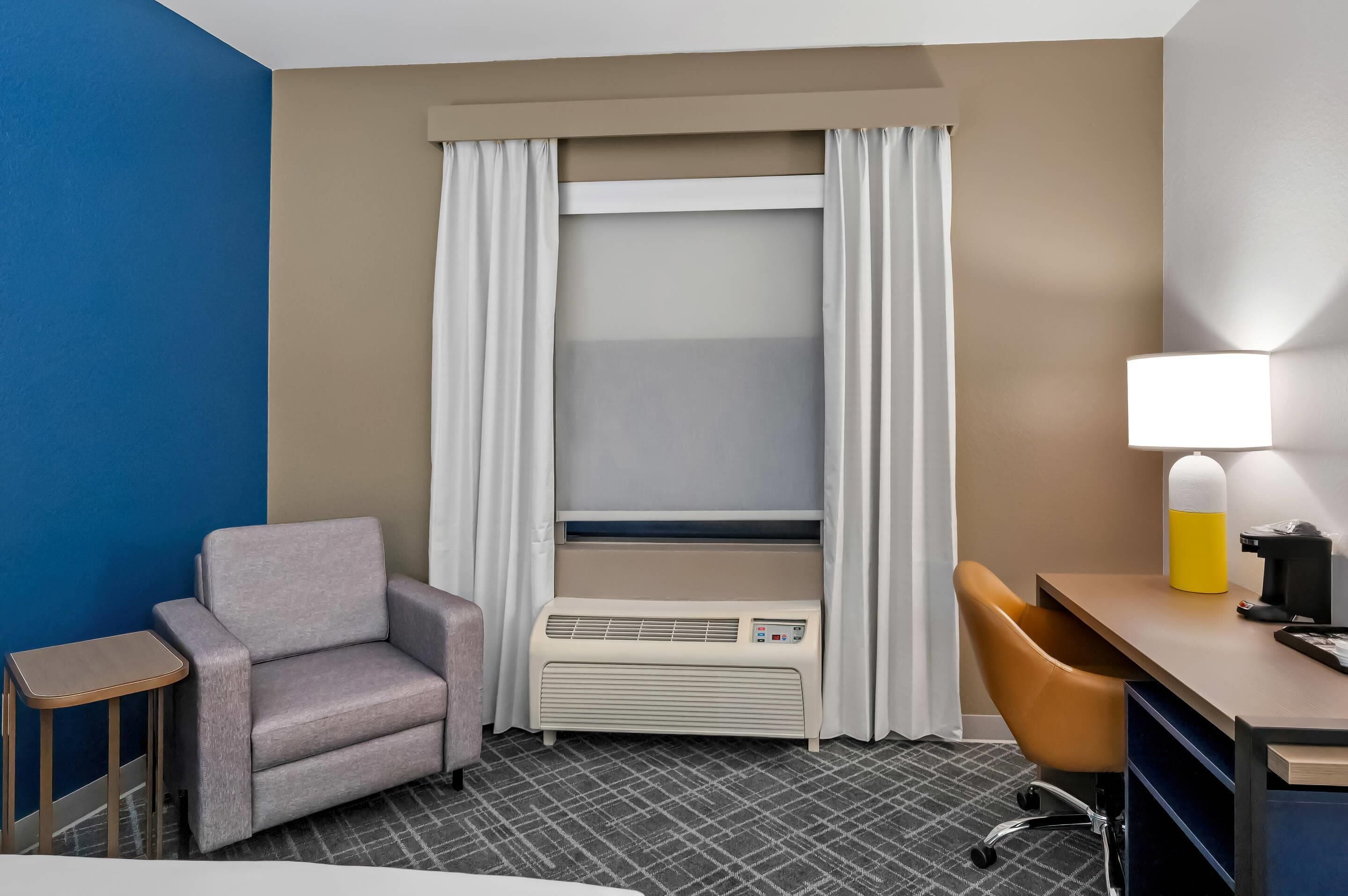 A hotel room with a bed, a desk, a chair, a small table, and a heater.