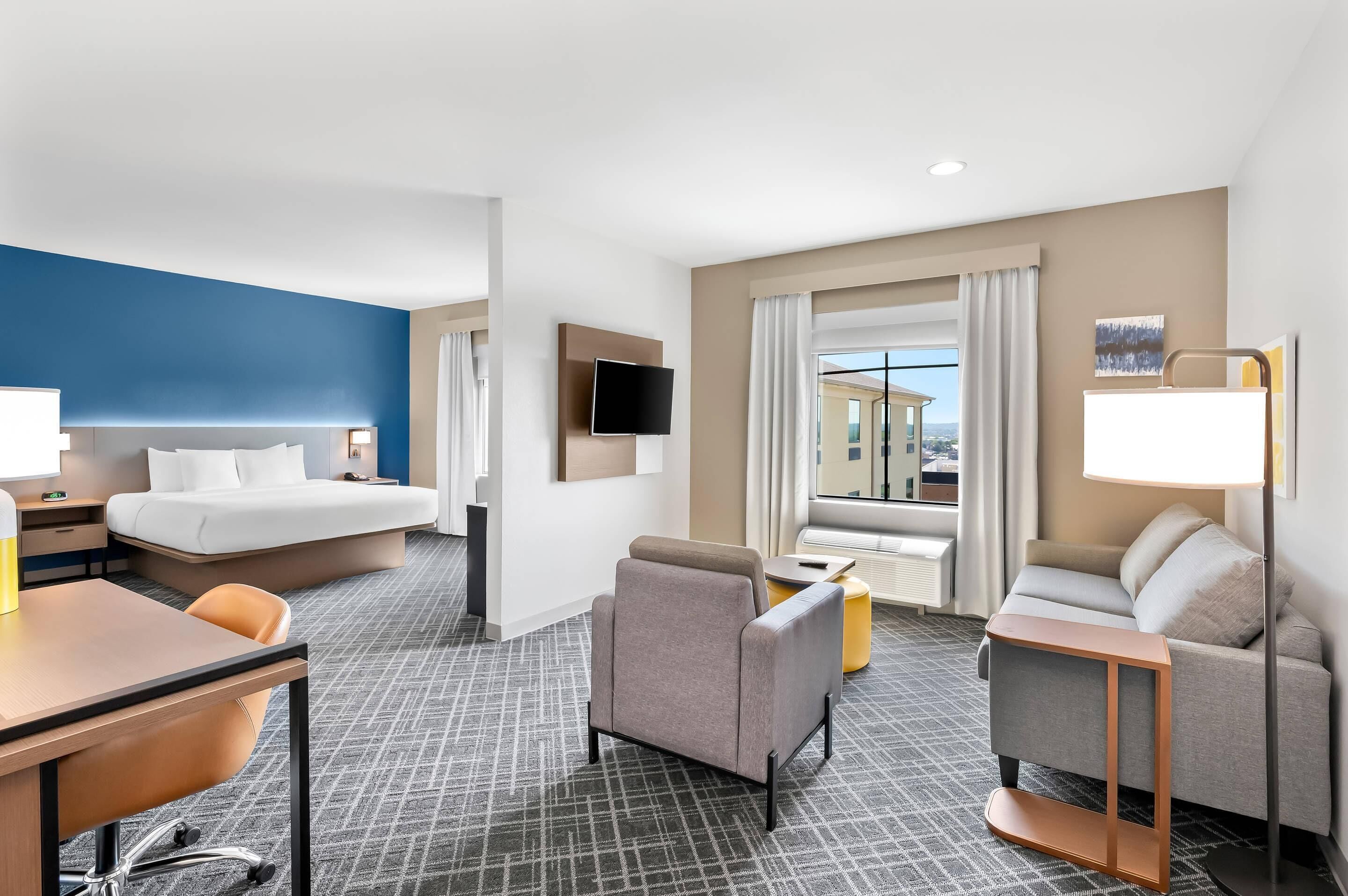 Habitación de hotel con cama, escritorio, silla, sofá, televisor, lámpara y ventana con vista a edificios.