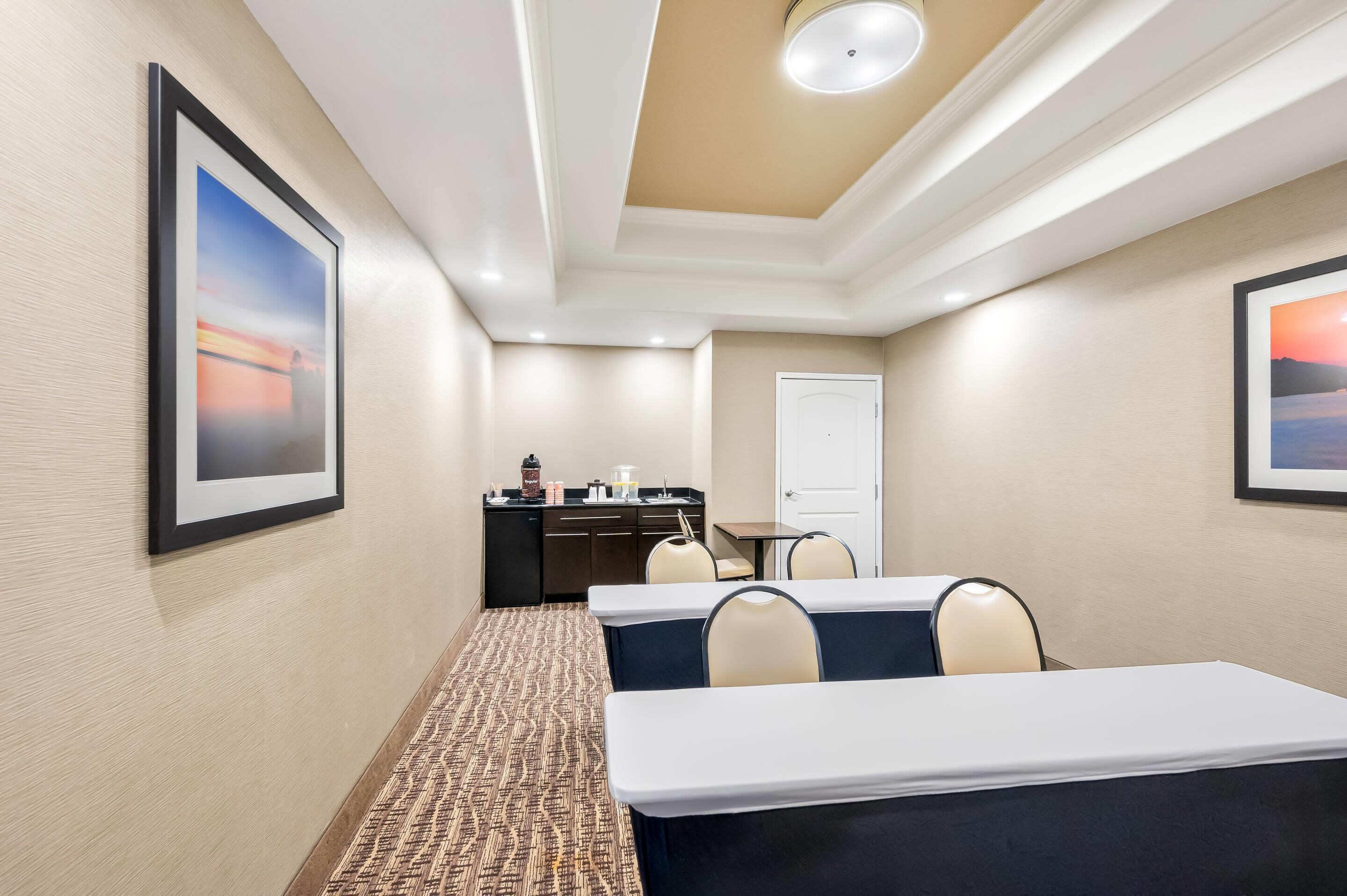 A hotel meeting room with two tables, chairs, a kitchenette, and framed artwork on the walls.
