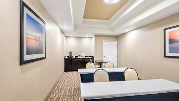 A hotel meeting room with two tables, chairs, a kitchenette, and framed artwork on the walls.